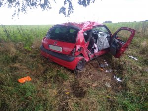 Na środku zdjęcia samochód osobowy Renault Clio koloru czerwonego z uszkodzeniami powypadkowym na prawej stronie. W tle pole.