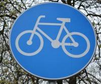 Znak informacyjny &quot;droga dla rowerów&quot;, okrągły, w niebieskim kolorze z rysunkiem roweru koloru białego. W tle gałęzie drzew.