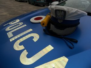 Na niebieskiej pokrywie silnika radiowozu widnieje biały napis Policja. Nad napisem leży czapka policjanta ruchu drogowego, tarcza do zatrzymywania pojazdów oraz urządzenie do sprawdzania stanu trzeźwości.