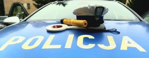 Niebieska pokrywa silnika radiowozy z białym napisem POLICJA. Na pokrywie leży biała czapka policjanta ruchu drogowego, żółte urządzenie AlcoBlow do sprawdzania stanu trzeźwości oraz biało- czerwona tarcza do zatrzymywania pojazdów, w tle szyba czołowa radiowozu.