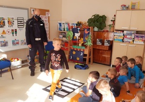 Sala przedszkolna. W prawej części zdjęcia grupa przedszkolaków siedzi na dywanie, po lewej stronie stoi policjant, na środku chłopiec przechodzący przez przejście dla pieszych wykonane z papieru.