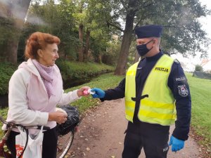 Po lewej stronie stoi starsza kobieta z rowerem, po prawej stronie stoi umundurowany policjant w kamizelce odblaskowej i wręcza kobiecie odblask. W tle aleja parkowa.