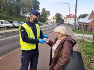 Po lewej stronie stoi umundurowany policjant w kamizelce odblaskowej i wręcza element odblaskowy kobiecie stojącej w prawej części fotografii. W tle ulica miasta.