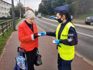 Po prawej stronie stoi umundurowana policjantka w kamizelce odblaskowej i wręcza element odblaskowy starszej kobiecie stojącej w lewej części fotografii. W tle ulica miasta.