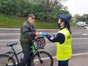 Po prawej stronie stoi umundurowana policjantka i wręcza element odblaskowy seniorowi na rowerze, który znajduje się w lewej części fotografii.