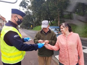 Po lewej stronie stoi policjant w kamizelce odblaskowej. Po prawej stronie stoją seniorzy: kobieta i mężczyzna, którym funkcjonariusz wręcza odblaski.