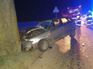 Pora nocna. na pierwszym planie samochód osobowy rozbity o drzewo znajdujące się w lewej części zdjęcia. Auto ma zbita szybę czołową oraz rozbity przód pojazdu. W tle wóz strażacki i strażacy.