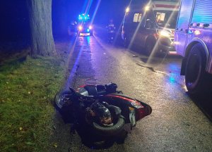 Pora nocna. Na pierwszym planie przewrócony motorower koloru czerwonego. Pojazd leży na jezdni. Po lewej stronie widać pobocze i drzewo, po prawej część wozu strażackiego, karetkę pogotowia, strażaka oraz kolejny pojazd uprzywilejowany z włączonymi sygnałami świetlnymi w kolorze niebieskim.