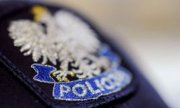 Fragment czapki służbowej od granatowego munduru. Biały orzeł w złotej koronie. Pod nim niebieska wstęga z napisem POLICJA.