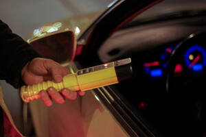 Pora nocna, na zdjęciu ujęty moment sprawdzania stanu trzeźwości. Od lewej strony ręka policjanta trzymająca żółte urządzenie do sprawdzania stanu trzeźwości.Prawa strona zdjęcia przedstawia fragment pojazdu z otwartą szybą lewą boczną.