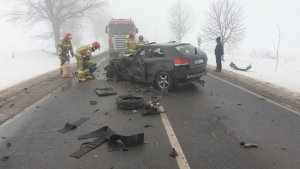 Miejsce zdarzenia drogowego. Silne zamglenie. Na pierwszym planie rozbity pojazd osobowy Audi koloru czarnego  z widocznymi uszkodzeniami bocznymi i przednimi. Wokół rozrzucone elementy auta. W tle ciężarówka z naczepą stojąca na całej szerokości jezdni. W oddali osoby obsługujące zdarzenie drogowe.