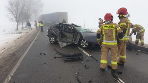 Pora dzienna, silne zamglenie. Na pierwszym planie strażacy i całkowicie zniszczony pojazd Audi stojący na środku jezdni, wokół rozrzucone drobne elementy pojazdu. W tle pojazd ciężarowy z naczepą stojący na całej szerokości jezdni. W różnych miejscach stoi kilka osób: strażacy policjant, inne osoby.