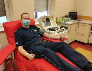 Policjant umundurowany w trakcie oddawania osocza. Funkcjonariusz siedzi na czerwonym fotelu w pomieszczeniu centrum krwiodawstwa, obok fotela stoi aparatura do pobierania osocza.