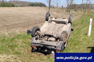Pojazd osobowy wywrócony kołami do góry leżący na trawiastym poboczu drogi. w tle pole, pora dzienna.