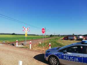 Pora dzienna. Przejazd kolejowy. Przed przejazdem w prawej częsci zdjecia stoi oznakowany policyjny radiowóz. W oddali straż pożarna i torowisko.