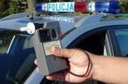 Na pierwszym planie prawa dłoń trzymająca alcosensor - urządzenie do badania stanu trzeźwości. W tle policyjny radiowóz oznakowany. Pora dzienna.