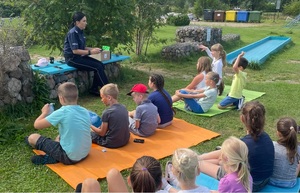 Pora dzienna. dzieci siedzą na kolorowych kocykach rozłożonych na trawie; w tle policjantka siedząca na ławeczce, w ręku trzyma granatową teczkę. w tle krzewy i pojemniki do segregacji śmieci.