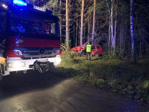 Na pierwszym planie fragment wozu strażackiego. W tle policjant w kamizelce odblaskowej z napisem policja stojący przed rozbitym autem z osobowym koloru czerwonego typ kombi. W tle las. Pora nocna.