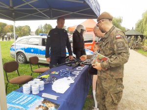 Pora dzienna. Stoisko Policji, na stołach gazety i gadżety policyjne oraz broń palna. Po prawej stronie żołnierz po prawej policjant, w tle radiowóz policyjny.