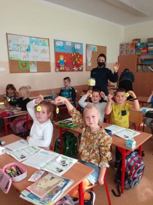 Sala lekcyjna. Dzieci siedzące w ławkach w rękach trzymają odblaski. W tle policjantka z odblaskiem w ręku.