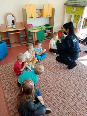 Sala zajęciowa. Dzieci siedzące na dywanie. Przed jednym dzieckiem kuca policjantka i wręcza odblask.