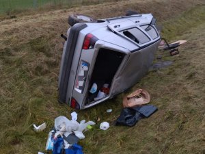 Pora dzienna. Pojazd osobowy koloru szarego leżący na boku. Posada uszkodzenia pokolizyjne. Przy nim, na trawie porozrzucane przedmioty. Pojazd leży w rtawiastym rowie.