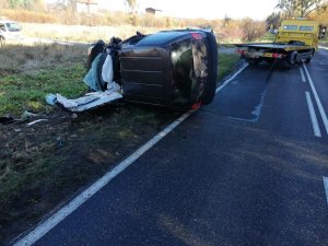 Pora dzienna. Miejsce zdarzenia drogowego. W lewej części fotografii przewrócone na bok rozbite auto leżące na poboczu. W centralnej i prawej części zdjęcia jest jezdnia. W tle żółta laweta.