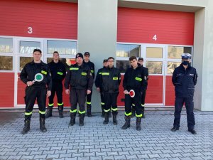 Pora dzienna. Po prawej stronie policjant ruchu drogowego . Po lewej i na środku grupa 8 druhów z ochotniczej straż pożarnej. W tle czerwone drzwi od garażu straży pożarnej.