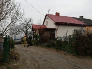 Pora dzienna. Brama wjazdowa na posesję. Po prawej stronie budynek jednorodzinny  z elewacją koloru kremowego i  czerwonym dachem. W głębi stoi radiowóz policyjny i strażak i mężczyzna ubrany w zielone moro.