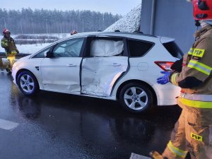 Por dzienna. Jezdnia a na niej strojący pojazd osobowy koloru białego z uszkodzonymi lewymi drzwiami. Prawej strony stoi strażak.