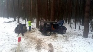 Pora dzienna. Las, na ziemi ścieg a w tle jezdnia. /Po prawej stronie samochód osobowy między drzewami, W pobliży przyczepka samochodowa i mężczyzna w odblaskowej, żółtej kurtce.