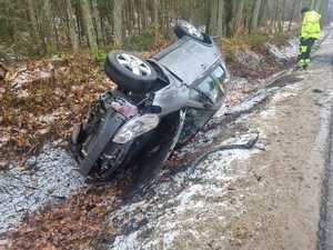 Pora dzienna. Droga prowadząca przez las. W rowie auto osobowe typu suv leżące na dachu. Widoczne uszkodzenia pokolizyjne.