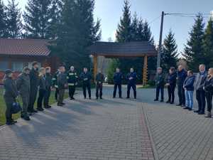 Pora dzienna. Uczestnicy wiosennej kampanii odnowieniowej stojący na placu w półokręgu. W tle budynek z drewna, drzewa i bezchmurne niebo.