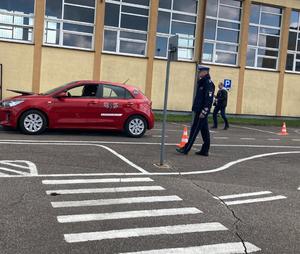 Powiatowy Turniej Motoryzacyjny. Sędziujący policjant w trakcie etapu praktycznego stojący na placu, po którym przemieszcza się pojazd osobowy koloru czerwonego.