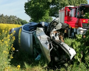 Pora dzienna, pobocze drogi. Na środku podwozie auta uczestniczącego w zdarzeniu drogowym. Po lewej stronie pole rzepaku, w tle wóz strażacki.