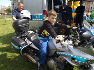 Chłopiec siedzący na motocyklu policyjnym. W tle policjanci i policyjny bus.