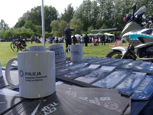 Pora dzienna. na pierwszym planie biały kubek z napisem Komenda Powiatowa Policji w Nidzicy, ulotki i gazety policyjne. W tle trawnik i uczestnicy festynu.