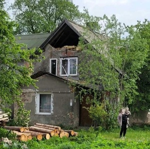 Pora dzienna Budynek mieszkalny z uszkodzonym szczytem. Przed budynkiem stoi kobieta z dzieckiem na ręku. Wokół trawa i drzewa.