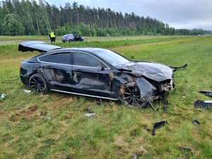 Pora dzienna, na prawie stoi rozbity samochód osobowy koloru czarnego. w tle na poboczu drogi stoi policjant i 2 auta osobowe. W głębi las.