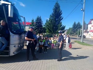 Policjanci ruchu drogowego. za nimi stoi grupa dzieci. po lewej astonie autobus. Pora dzienna,