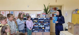 Sala lekcyjna grupa dzieci stoi po lewej stronie, po prawej stronie policjantka trzymająca książkę Na ratunek Poli.