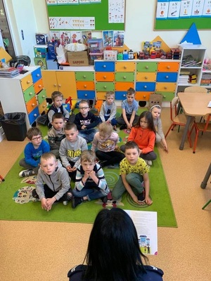 Sala lekcyjna, grupa dzieci siedzi na dywanie i słucha bajkę czytaną przez policjantkę.