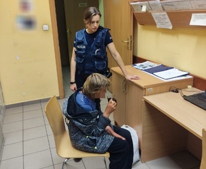 Pomieszczenie. Starsza kobieta siedzi na krześle i pije kawę. Przy niej stoi policjantka.