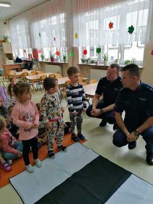 Sala zajęciowa. Na podłodze mata imitująca przejście dla pieszych. Wokół niej stojące dzieci i dwóch kucających policjantów.