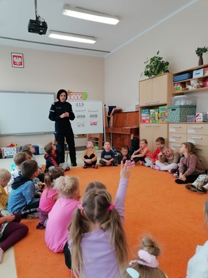 Sala przedszkolna. Grupa dzieci siedząca na pomarańczowym dywanie, przed nimi  stojąca policjantka.