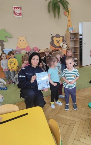 Sala przedszkolna. Policjantka kuca i trzyma dyplom, który otrzymała od dzieci. Obok niej stoi grupa dzieci.