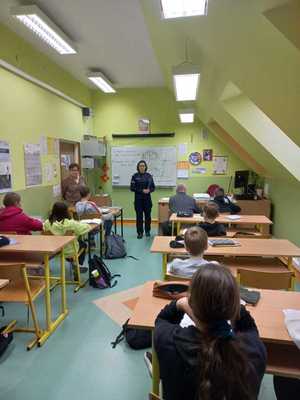 Klasa lekcyjna. Dzieci siedza w ławkach. Przed nimi stoi policjantka wygłaszająca prelekcję.