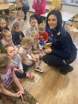 Sala przedszkolna. Policjantka kuca przy grupie dzieci. w rękach trzyma odblaski w kształcie misia.