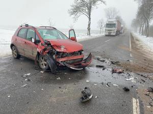 Droga. Na niej rozbite auto osobowe koloru czerwonego. Zniszczony przód auta. W tle ciężarówka i drzewa rosnące przy poboczu drogi.