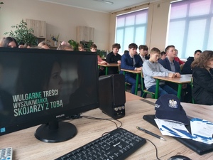 Klasa szkolna. Biurko a na nim monitor komputera, klawiatura, czapka policyjna, ulotki. W tle dzieci siedzące w ławkach.